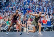 sivasangari - Wilayah.com.my Two female squash players sprint toward the ball on a crowded arena court, racquets raised, mid-match action amid spectators.
