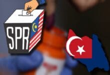 Person in a blue glove casts a ballot into an SPR box featuring a Malaysian flag motif; a red Turkish map with white crescent and star is nearby.