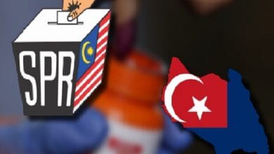 Person in a blue glove casts a ballot into an SPR box featuring a Malaysian flag motif; a red Turkish map with white crescent and star is nearby.