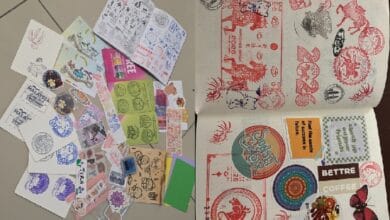 stamp 2 - Wilayah.com.my A collection of decorative stamps, stickers, and cutouts spread across a tiled floor. On the right, an open notebook shows stamp designs and colorful sticker sheets on the pages.