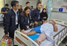 Medical team and visitors stand beside a patient in a hospital bed, the patient’s left leg in a cast and bandages visible.