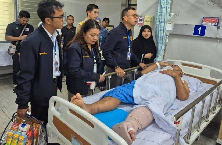 Medical team and visitors stand beside a patient in a hospital bed, the patient’s left leg in a cast and bandages visible.