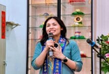 Smiling woman in a turquoise top and blue ceremonial scarf speaks into a handheld microphone, with glass display shelves and colorful decorations behind her.