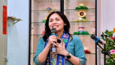 Smiling woman in a turquoise top and blue ceremonial scarf speaks into a handheld microphone, with glass display shelves and colorful decorations behind her.