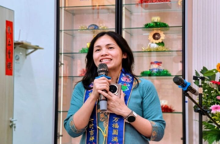 Smiling woman in a turquoise top and blue ceremonial scarf speaks into a handheld microphone, with glass display shelves and colorful decorations behind her.