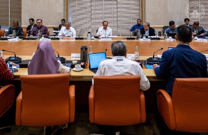 Officials sit around a long conference table in a formal meeting, viewed from behind three orange chairs in the foreground.
