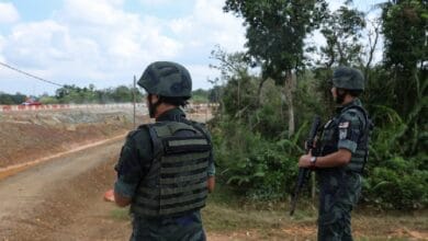 Two soldiers in camouflage uniforms and helmets stand on a dirt road near trees, armed with rifles, surveying the area.