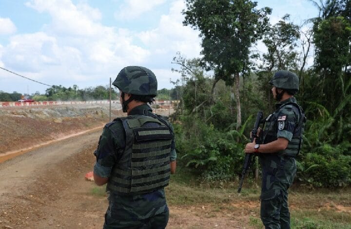 Two soldiers in camouflage uniforms and helmets stand on a dirt road near trees, armed with rifles, surveying the area.