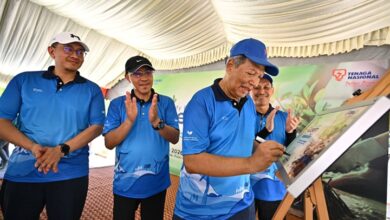 tnb - Wilayah.com.my Men in blue polo shirts under a tent watch one man sign a board at an outdoor event.