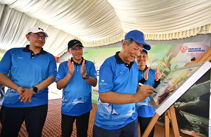 Men in blue polo shirts under a tent watch one man sign a board at an outdoor event.