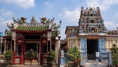 tokong kuil - Wilayah.com.my Ornate temple entrance with red gate and green tiled roof, flanked by potted plants, across from a blue‑and‑gold pagoda‑style shrine.