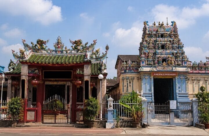 tokong kuil - Wilayah.com.my Ornate temple entrance with red gate and green tiled roof, flanked by potted plants, across from a blue‑and‑gold pagoda‑style shrine.