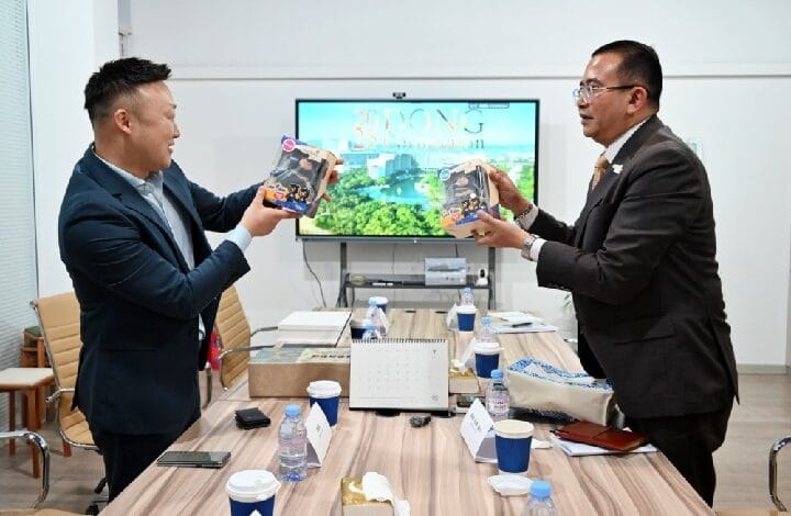 Two businessmen in suits exchange boxed products across a conference table in a meeting room, smiling.