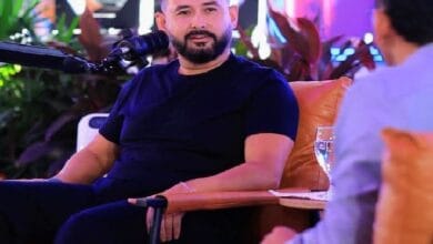 tunku mahkota ismail - Wilayah.com.my Bearded man in a dark navy T-shirt sits on an orange chair during a talk, microphone in front of him, conversing with a person off to the right in a bright setting.