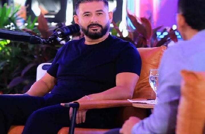tunku mahkota ismail - Wilayah.com.my Bearded man in a dark navy T-shirt sits on an orange chair during a talk, microphone in front of him, conversing with a person off to the right in a bright setting.