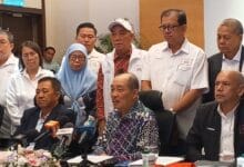 upko grs - Wilayah.com.my Group of officials and reporters at a press conference around a table with multiple microphones, some seated and others standing in white uniforms and formal attire.