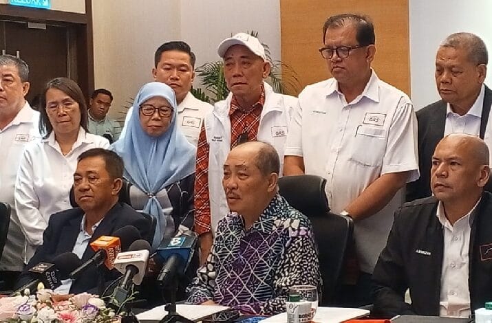 Group of officials and reporters at a press conference around a table with multiple microphones, some seated and others standing in white uniforms and formal attire.