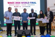 Six people on a stage at a media launch for the WAGC 2026 World Final, holding promotional signs for Malaysia and Visit Johor, with a large screen behind and confetti on the floor.
