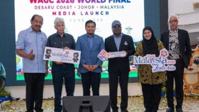 Six people on a stage at a media launch for the WAGC 2026 World Final, holding promotional signs for Malaysia and Visit Johor, with a large screen behind and confetti on the floor.