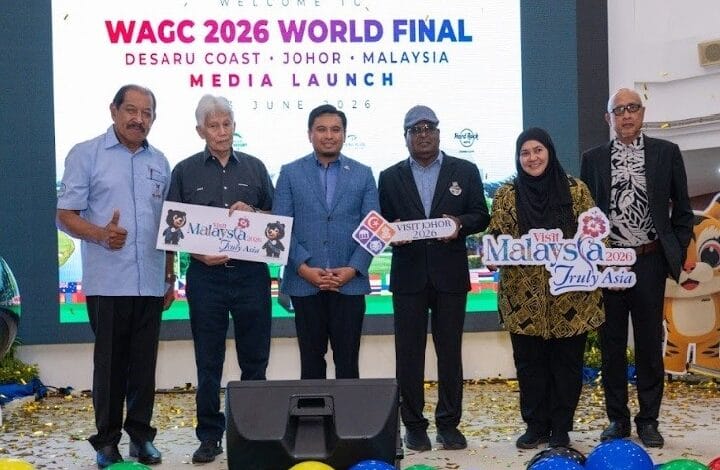 Six people on a stage at a media launch for the WAGC 2026 World Final, holding promotional signs for Malaysia and Visit Johor, with a large screen behind and confetti on the floor.