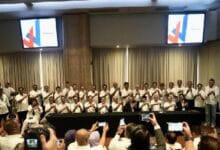 wawasan - Wilayah.com.my Group of people in white shirts posing for a group photo at a conference, behind a long table with cameras in the foreground. Screens with logos shown in the background.