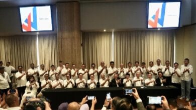 Group of people in white shirts posing for a group photo at a conference, behind a long table with cameras in the foreground. Screens with logos shown in the background.