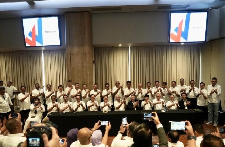 wawasan - Wilayah.com.my Group of people in white shirts posing for a group photo at a conference, behind a long table with cameras in the foreground. Screens with logos shown in the background.