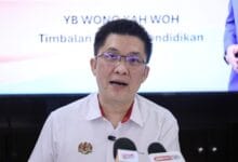 wong kah woh - Wilayah.com.my Man in a white shirt speaking at a press conference, microphones in front, with blurred backdrop showing text in a formal setting.
