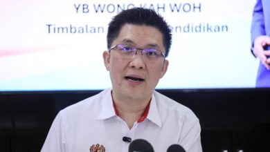 wong kah woh - Wilayah.com.my Man in a white shirt speaking at a press conference, microphones in front, with blurred backdrop showing text in a formal setting.