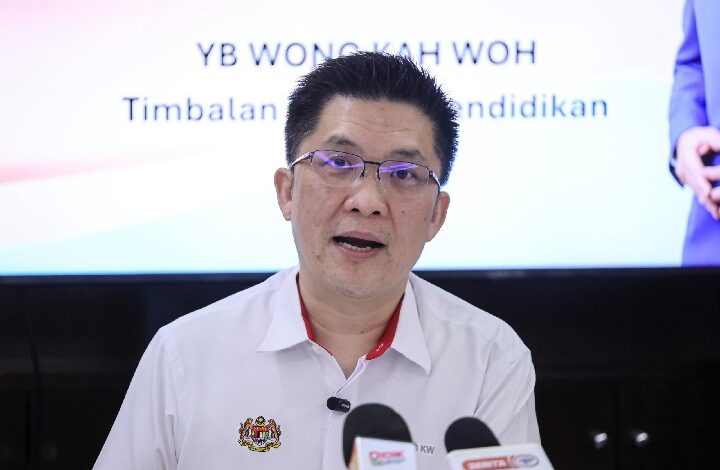 wong kah woh - Wilayah.com.my Man in a white shirt speaking at a press conference, microphones in front, with blurred backdrop showing text in a formal setting.