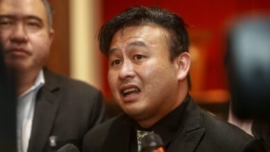 Asian man in suit speaking to reporters, microphones in foreground at a press event