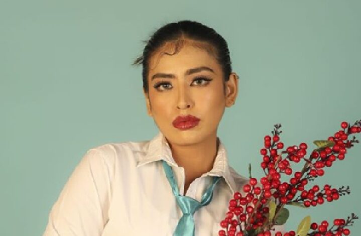 yazmin aziz - Wilayah.com.my Portrait of a woman in a white shirt and turquoise tie, posing with a red berry branch against a mint backdrop.