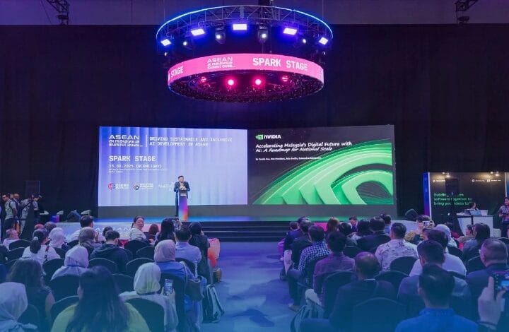 Presenter on stage at a conference with large screens and an audience seated in front, blue circular lighting overhead.
