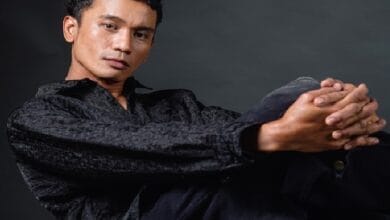 Portrait of a man seated against a dark backdrop, wearing a black textured shirt with hands clasped in front.