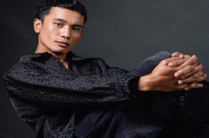Portrait of a man seated against a dark backdrop, wearing a black textured shirt with hands clasped in front.