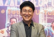 zizan nin - Wilayah.com.my Smiling person in a gray blazer, black shirt, round glasses and black beret, posing in front of a purple neon-backed event backdrop.
