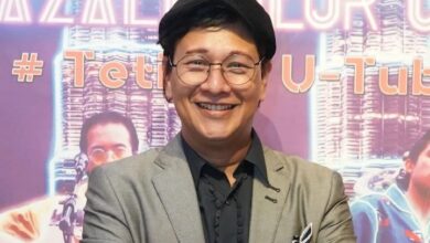zizan nin - Wilayah.com.my Smiling person in a gray blazer, black shirt, round glasses and black beret, posing in front of a purple neon-backed event backdrop.