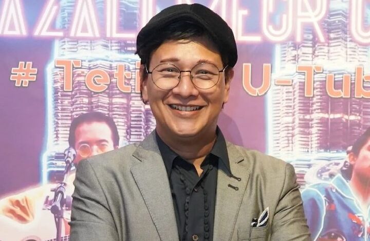 zizan nin - Wilayah.com.my Smiling person in a gray blazer, black shirt, round glasses and black beret, posing in front of a purple neon-backed event backdrop.