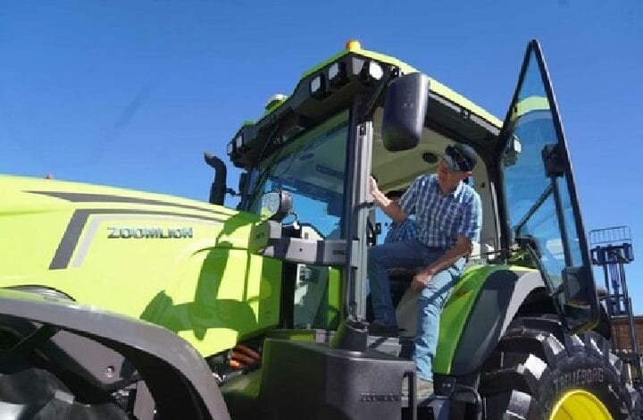 Man in a plaid shirt climbing into a bright green agricultural tractor with open door against a clear blue sky.