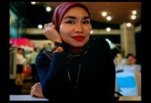 adzrin adzhar 2 - Wilayah.com.my Portrait of a woman wearing a red striped hijab and dark top, smiling with her chin resting on her hand in a warm indoor setting.