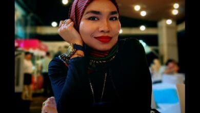adzrin adzhar 2 - Wilayah.com.my Portrait of a woman wearing a red striped hijab and dark top, smiling with her chin resting on her hand in a warm indoor setting.