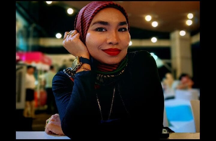 adzrin adzhar 2 - Wilayah.com.my Portrait of a woman wearing a red striped hijab and dark top, smiling with her chin resting on her hand in a warm indoor setting.