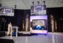 ampera 2 - Wilayah.com.my Speaker at a podium presenting beside a large futuristic capsule on a stage, with pyrotechnics and tall screens above.