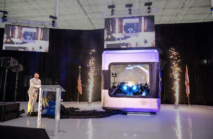 ampera 2 - Wilayah.com.my Speaker at a podium presenting beside a large futuristic capsule on a stage, with pyrotechnics and tall screens above.