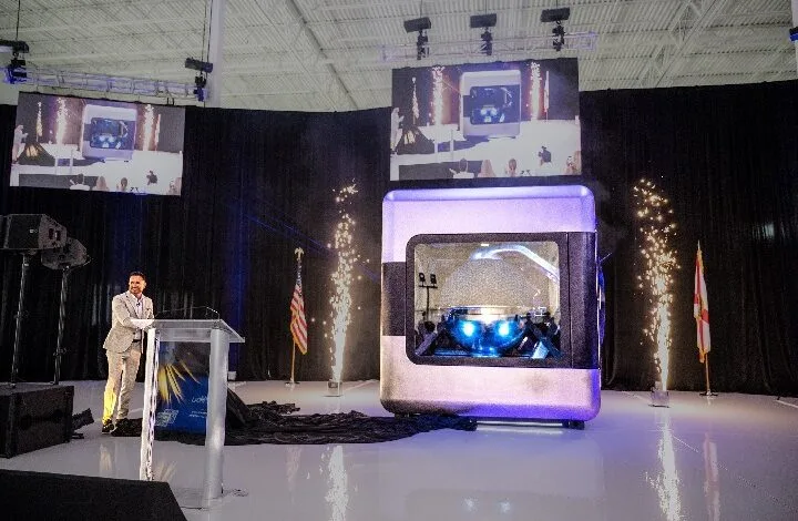 ampera 2 - Wilayah.com.my Speaker at a podium presenting beside a large futuristic capsule on a stage, with pyrotechnics and tall screens above.