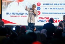 anwar - Wilayah.com.my Man on stage with microphone addressing a crowd at a campaign event, with a large screen displaying event details in the background.