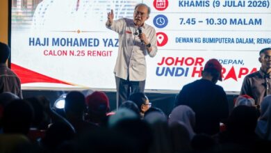 anwar - Wilayah.com.my Man on stage with microphone addressing a crowd at a campaign event, with a large screen displaying event details in the background.