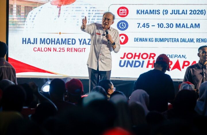 anwar - Wilayah.com.my Man on stage with microphone addressing a crowd at a campaign event, with a large screen displaying event details in the background.