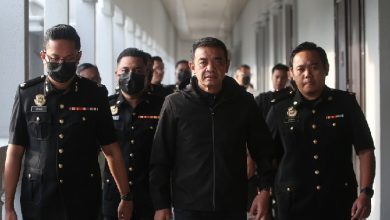 Man in a black jacket walks ahead as uniformed officers flank him down a hallway, some wearing masks.