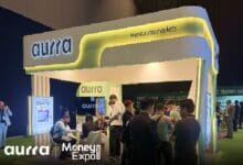 aurra 2 - Wilayah.com.my Aurra Markets booth at a Money Expo with a curved, yellow-lit display and people networking nearby.